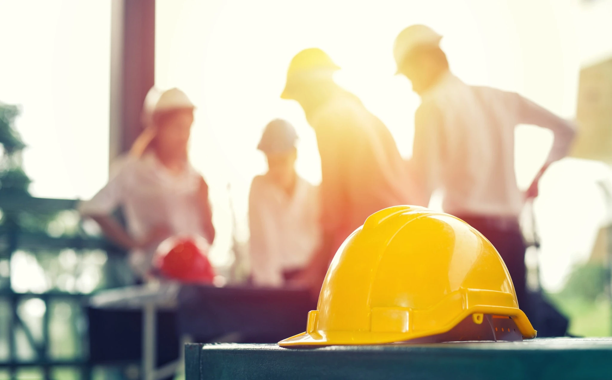 A yellow hard hat in focus with a blurred team of safety professionals meeting on-site in the background.